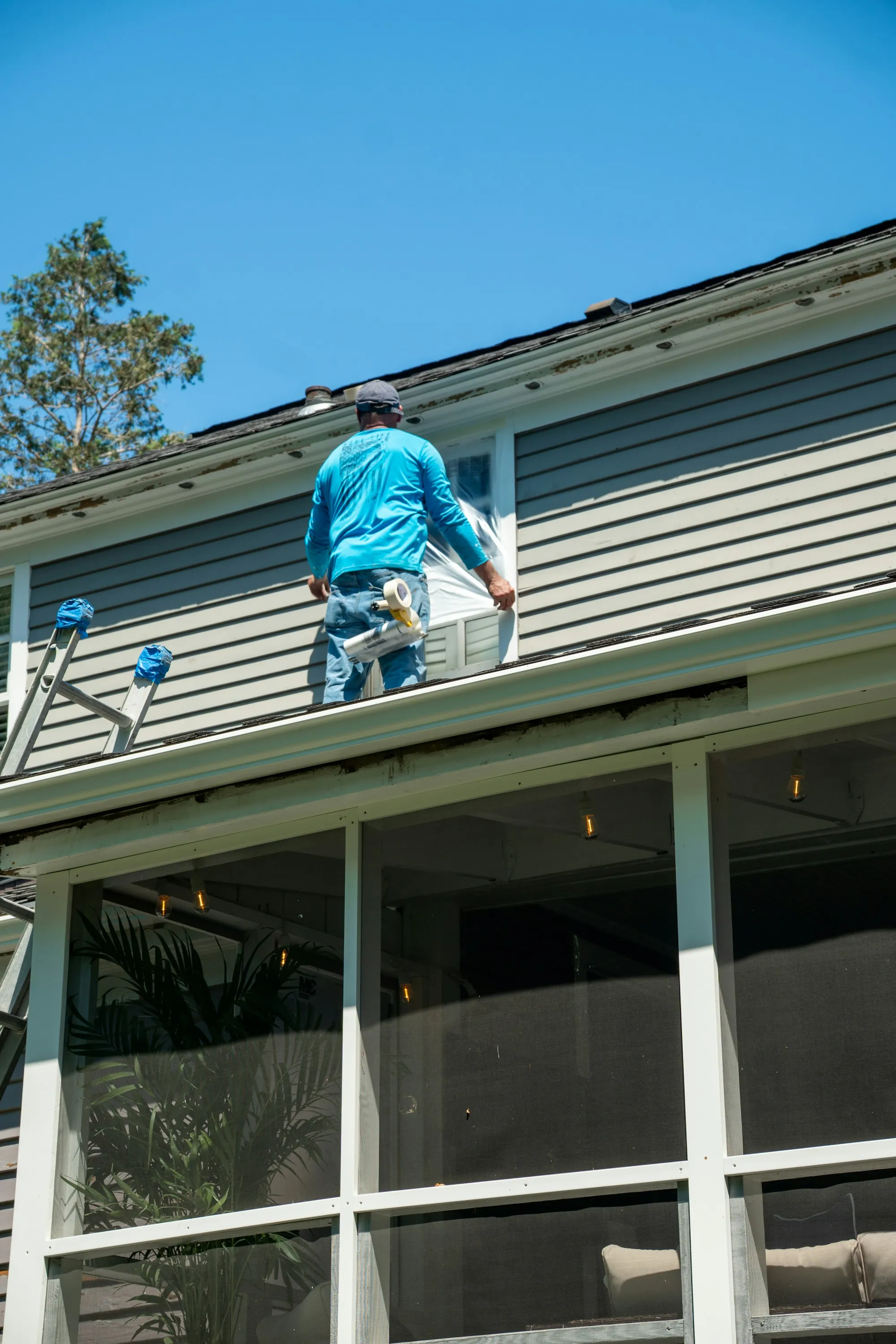Worker on a lower roof taping plastic over a window on a sided house